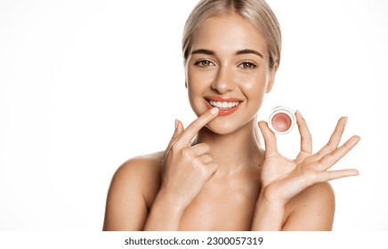 Close up of blond smiling beauty model, girl applies lip balm, pink gloss for natural shine and glow, uses makeup on clean, perfect skin without blemishes, white background.