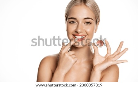 Close up of blond smiling beauty model, girl applies lip balm, pink gloss for natural shine and glow, uses makeup on clean, perfect skin without blemishes, white background.