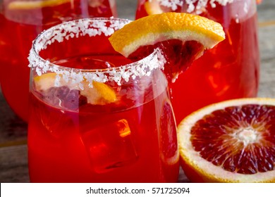 Close Up Of Blood Orange Margaritas With Margarita Salt On Rim Garnished With Fresh Blood Orange Slice And Fruit Half Sitting Table