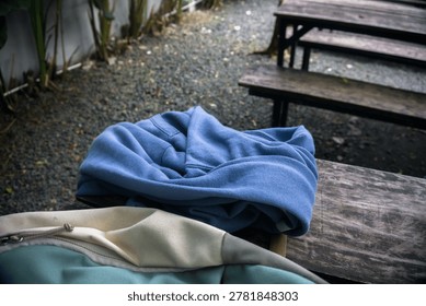 close up of a blue sweater and a backpack placed on a rustic wooden bench in an outdoor cafe garden with gravel ground portraying a relaxing leisure break atmosphere