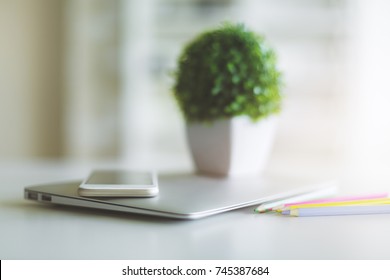 Close Up Of Blurry Office Desk Top With Laptop, Smartphone, Colorful Pencils And Decorative Green Plant