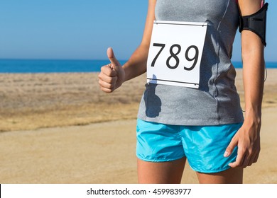 Close up body part of female athlete with race start number doing thumbs up outdoors.