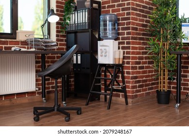 Close up of bookshelf and water dispenser in business office. Empty desk with decorations and watercooler in startup space, shelves with minimal decor. Decorative plants and refreshment.