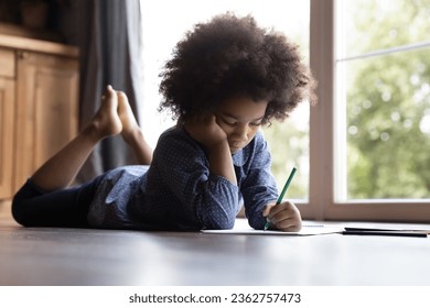 Close up bored African American little girl drawing and colorful pencils  lying warm wooden floor at home alone  adorable 7s child kid engaged in creative activity  spending leisure time