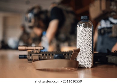 Close up of bottle full of plastic bullets and ammo on a table with airsoft players in a blurry background. Airsoft ammunition on table in bottle with sniper rifle next to it. War simulation game.