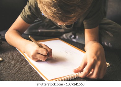 Close up bottom view young boy drawing face white sheet  Kid hold pencil   draw something  warm orange light at home  Children writing paper  Teen drawing sitting sofà