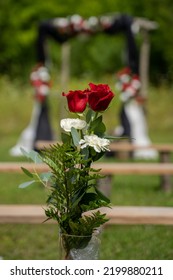 Close Up Of A Bouquet Of Two Red Roses And Two White Carnations At An Outdoor Wedding In Front Of An Arbor Decorated With Red And White Roses.