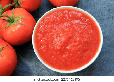 close up of bowl with tomato puree and some tomatoes next to it on gray table