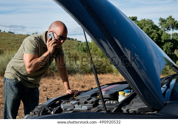 Close up of a broken down\
car, engine open, in a rural area and the driver looking at the\
engine
