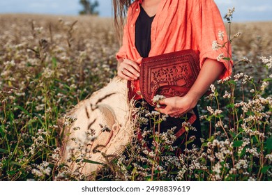 Close up of brown leather purse and straw hat in female hands. Woman holding accessories in blooming buckweat field among flowers at sunset. Boho style handbag - Powered by Shutterstock