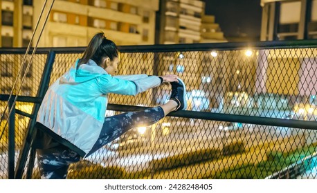 Close up of brunette female runner stretching her legs over bridge railing before training on city at night. Woman athlete warming up on evening in town. Health and fitness concept in urban setting. - Powered by Shutterstock