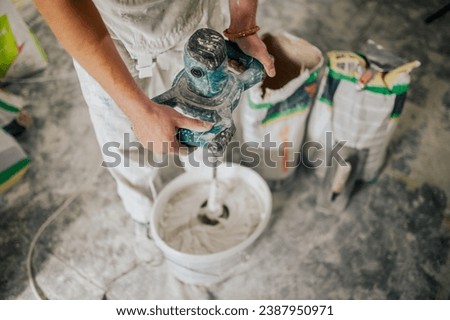 Close up of builder's hands mixing plaster with manual cement mixer while standing in dusty and messy room in renovation process. A constructor is preparing plaster for skim coating walls.