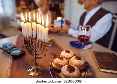 Close Up Of Burning Candles In Menorah With Mature Jewish Man Giving  Present To His Wife In The Background.