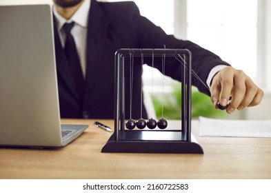 Close up of business leader playing with Newton's cradle on office desk with notebook PC. Man moves black pendulum ball, it hits others, and kinetic energy is transferred into potential and vice versa - Shutterstock ID 2160722583