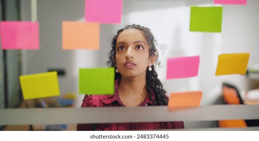 Close up of business woman standing indoor read new project sticky colorful notes puts day reminder stickers office glass board. Indian gen z curly hair female check ideas plan on memo paper job place - Powered by Shutterstock