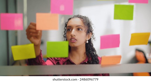 Close up of business woman standing indoor read new project sticky colorful notes puts day reminder stickers office glass board. Indian gen z curly hair female check ideas plan on memo paper job place - Powered by Shutterstock