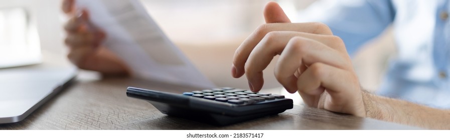 Close up businessman checking bills, holding receipt, using calculator, accountant calculating company expenses, financial report, sitting at wooden work desk, young male planning budget
