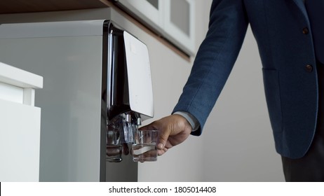 Close Up Of Businessman Hand Pouring Water From Cooler In Kitchen. Office Employee Using Water Cooler To Drink In Cafeteria. Male Hand In Suit Holding Glass Getting Water From Cooler