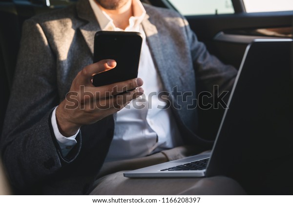 Close up of businessman\
holding mobile phone while sitting with laptop computer at the back\
of a car