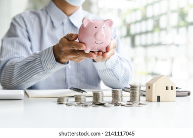 Close Up Businessman Holding Piggy Bank And Raw Coin With Mini Wood House.