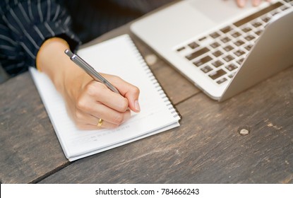 close up businesswoman hand writing content or somethings on notebook with using laptop at wooden table outside home , lifestyle concept