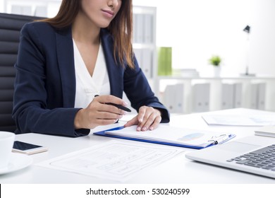 Close up of a businesswoman with tanned skin holding a pen and sitting at her table with a clipboard.