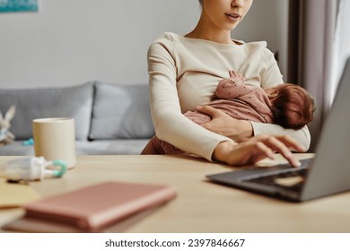 Close up of busy mother holding her baby while sitting at table and working on laptop at home, copy space - Powered by Shutterstock