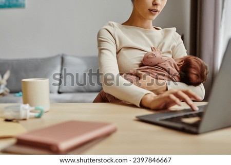 Close up of busy mother holding her baby while sitting at table and working on laptop at home, copy space