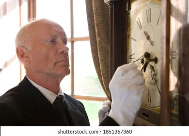 Close up of a butler adjusting the hands of an old grandfather clock on a sunny day inside a luxurious house