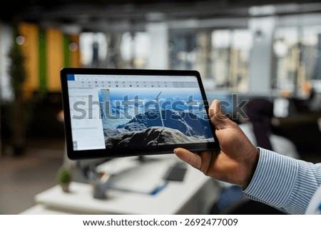 Close up of CAD engineer using tablet app in office to upgrade turbine, boosting power output, reducing carbon footprint. Worker using digital device to review windmill data to optimize clean energy