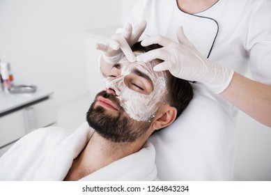 Close up of calm bearded man relaxing in beauty salon and hands in gloves touching his forehead while putting white clay on it
