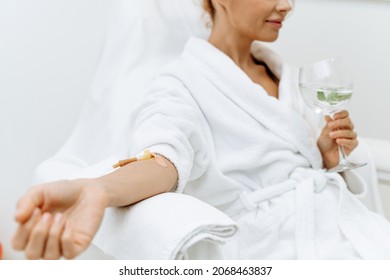 Close up of calm female patient sitting with tube and needle during IV infusion. Girl holding glass with lemon water and relaxing 