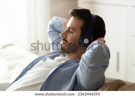 Close up calm peaceful man wearing headphones enjoying music, leaning back on cozy sofa with closed eyes, listening to pleasant sounds, daydreaming or napping, spending leisure time at home