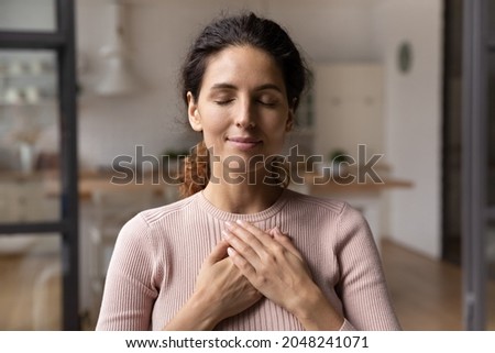 Close up of calm young Caucasian woman hold hands on heart chest feel grateful and thankful. Happy millennial female show gratitude, love and care, pray or visualize. Religion, faith concept.