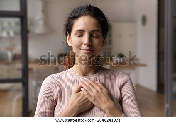 Close up of calm young Caucasian woman hold\
hands on heart chest feel grateful and thankful. Happy millennial\
female show gratitude, love and care, pray or visualize. Religion,\
faith concept.