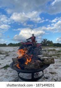 Close Up Of A Camp Site Fire With An ATV Rider On His Bike In The Background, Blurry, On Purpose.