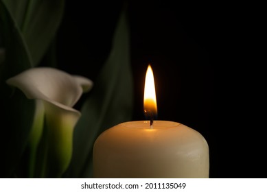 Close Up Of Candle Burning Flame In The Darkness And Arum Lilies Illuminated By The Candlelight Alongside In A Conceptual Image