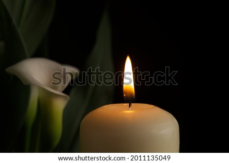 Close up of candle burning flame in the darkness and arum lilies illuminated by the candlelight alongside in a conceptual image