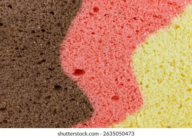 Close up captures the textured surface of a three layered sponge cake. The distinct colors a rich brown, a warm pink, and a bright yellow merge with soft, undulating lines.