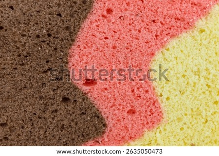 Close up captures the textured surface of a three layered sponge cake. The distinct colors a rich brown, a warm pink, and a bright yellow merge with soft, undulating lines.