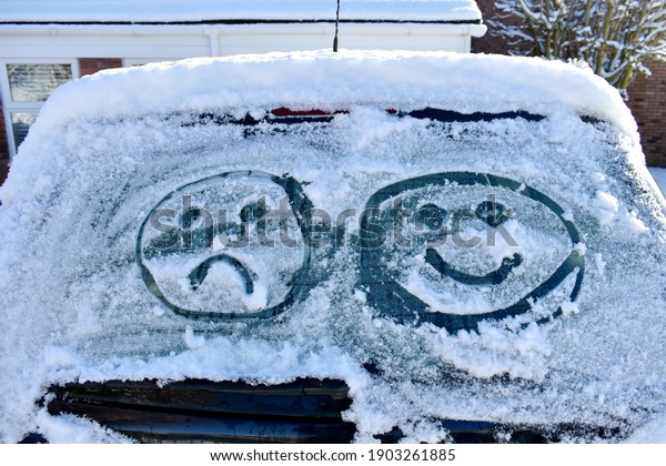 close up of car screen covered in snow with two emoji\
faces drawn into ice, one happy and one sad. outside on a cold\
sunny winter\'s day