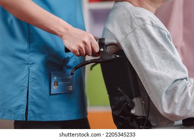 Close Up Of Caregiver Nurse Support Senior Elderly Man On Wheelchair In Living Room. Nurse Female Take Care Of Mature Patient Doing Physical Therapy At Nursing Home. Health Care Concept.