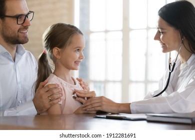 Close Up Of Caring Smiling Female Cardiologist Listen To Small Girl Child Patient Heart At Hospital Visit With Dad. Woman Doctor Or Nurse Examine Little Kid At Clinic Consultation. Healthcare Concept.