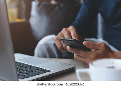 Close up of casual man or freelancer working on laptop computer and holding mobile smart phone with cup of coffee on table in coffee shop or cafe, working from cafe, freelance working concept.