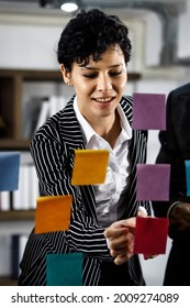 Close Up To Caucasian Beautiful Fashional Formal Businesswoman Writing And Brainstorming Creative Marketing Ideas On Colorful Memo Or Post It At Mirror With Mixed Race African Black Colleagues