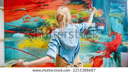 Close up of Caucasian cheerful happy young female artist in apron working in art studio painting using paint brush on big canvas making emotional colorful abstract masterpiece. Modern art concept