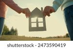 Close up Caucasian family woman man hands together holding cardboard house home model picture outdoors rural meadow field sunshine. Dream rent new flat real estate apartment selling property dwelling