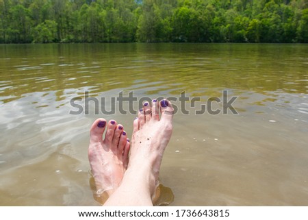 close up of caucasian feet with purple painted toe nails crossed at the ankle in brown and green lake water with tree and sky country background