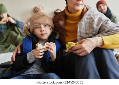 Close Up Of Caucasian Little Boy With Family In Refugee Shelter Eating Sandwich And Looking At Camera
