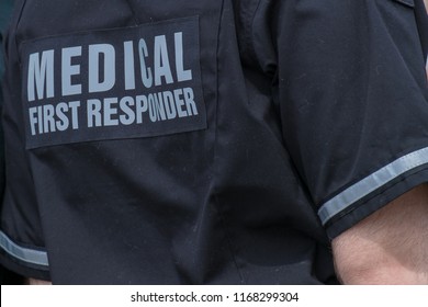 A close up of a caucasian male emergency health medical first responder or paramedic wearing a black uniform with grey letters. The ambulance attendant is wearing a short sleeve shirt with lettering.